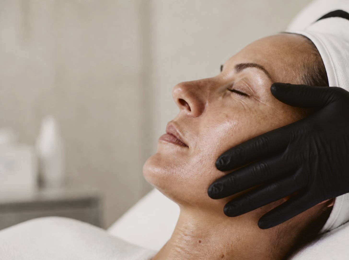 A woman relaxing during a skin treatment as a therapist rests gloved hands on her face